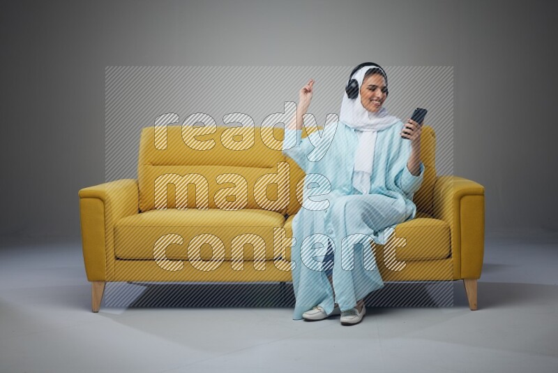A Saudi woman wearing a light blue Abaya and a white head scarf sitting on a yellow sofa and holding an iPad while wearing headphone eye level on a grey background