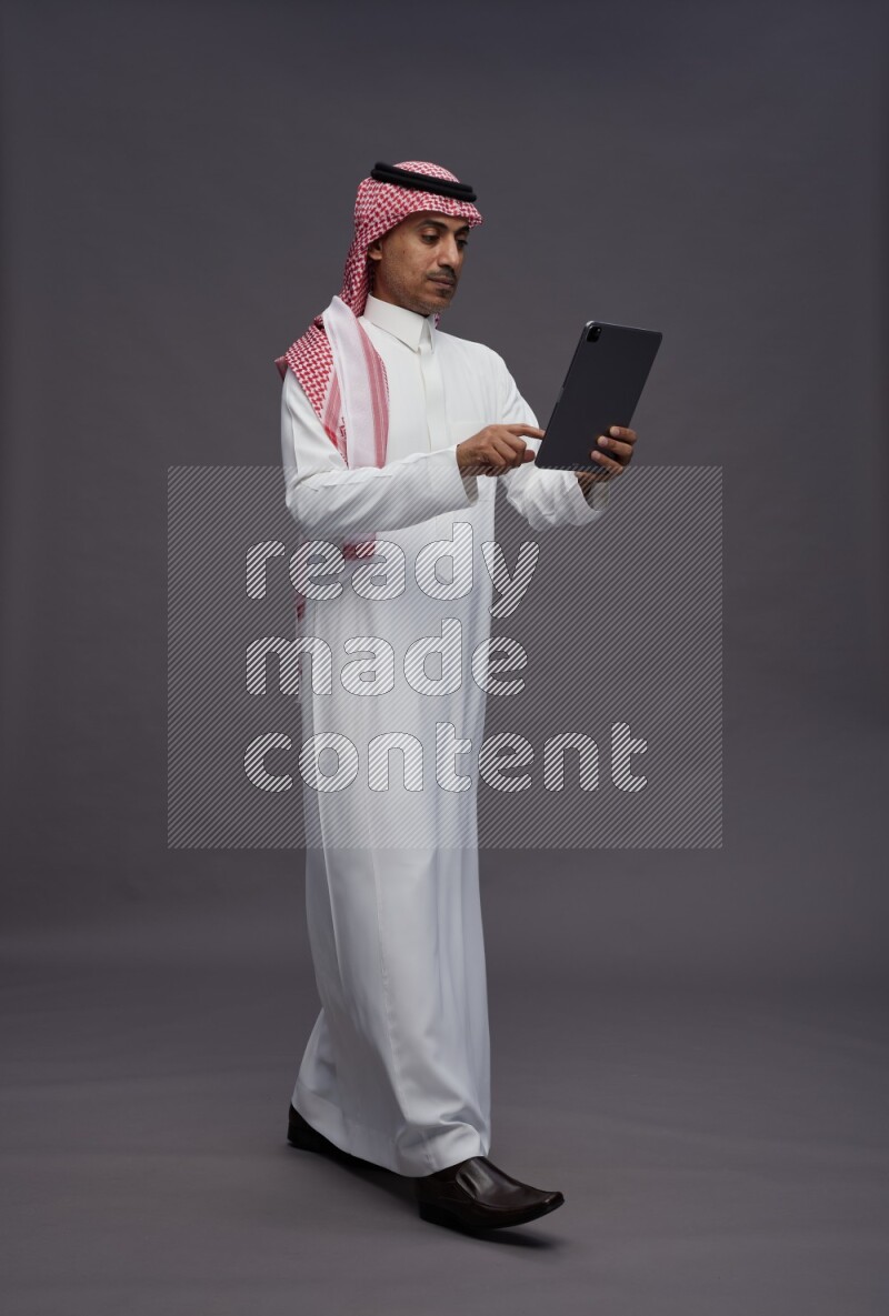 Saudi man wearing thob and shomag standing working on tablet on gray background
