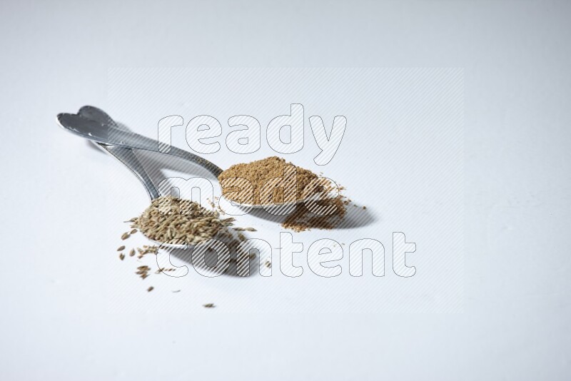 2 metal spoons full of cumin seeds and powder on white flooring