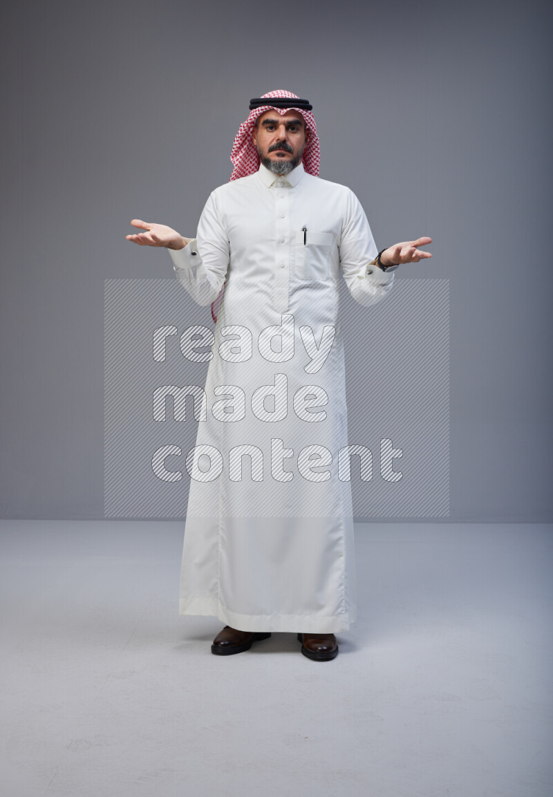 Saudi man Wearing Thob and red white Shomag standing interacting with the camera on Gray background