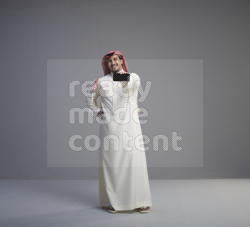 رجل سعودي يرتدي ثوب ابيض وشماغ احمر يشير الى هاتفه باتجاه الكاميرا