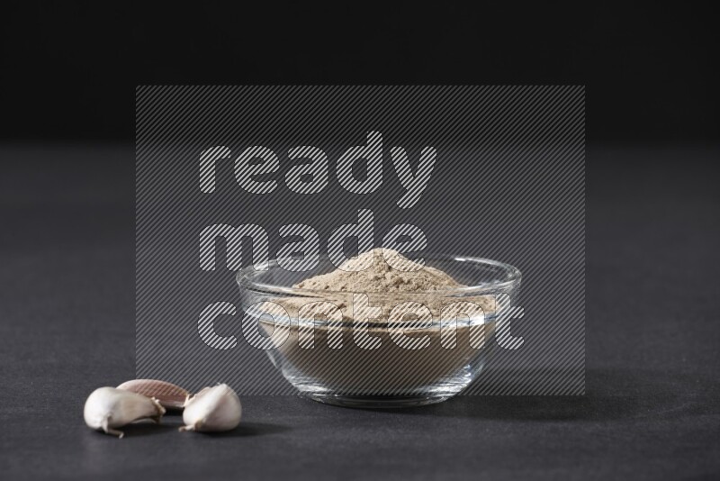 A glass bowl full of garlic powder with garlic bulb and some cloves beside it on a black flooring