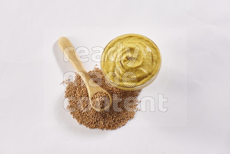 A glass bowl full of mustard paste with some of mustard seeds underneath it and a wooden spoon on white flooring