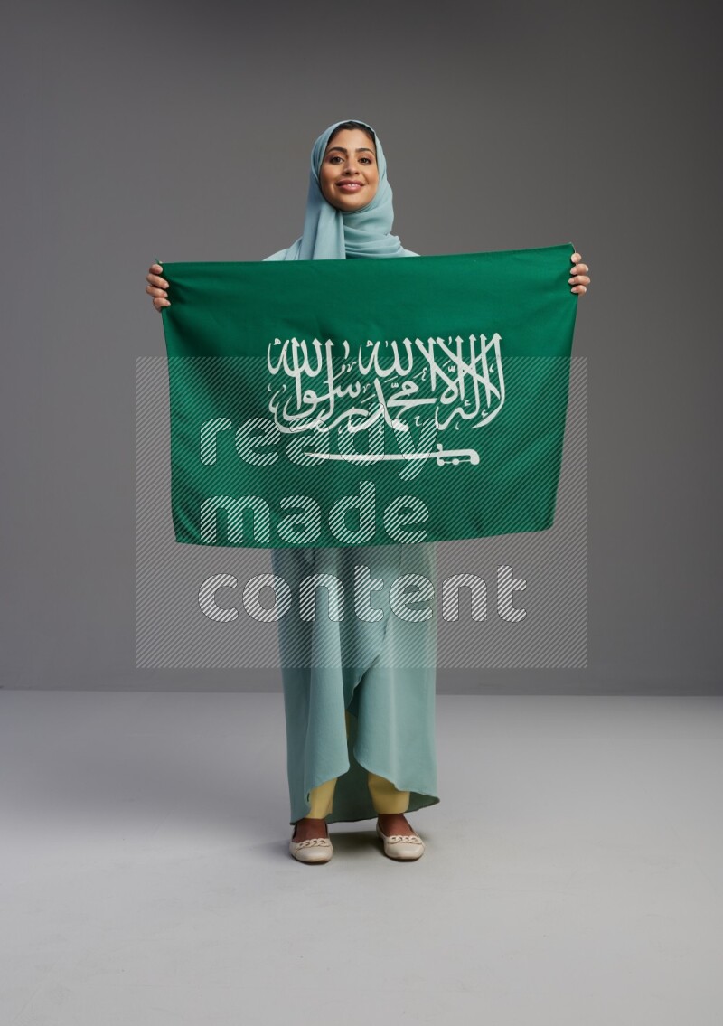 Saudi Woman wearing Abaya standing holding Saudi flag on Gray background