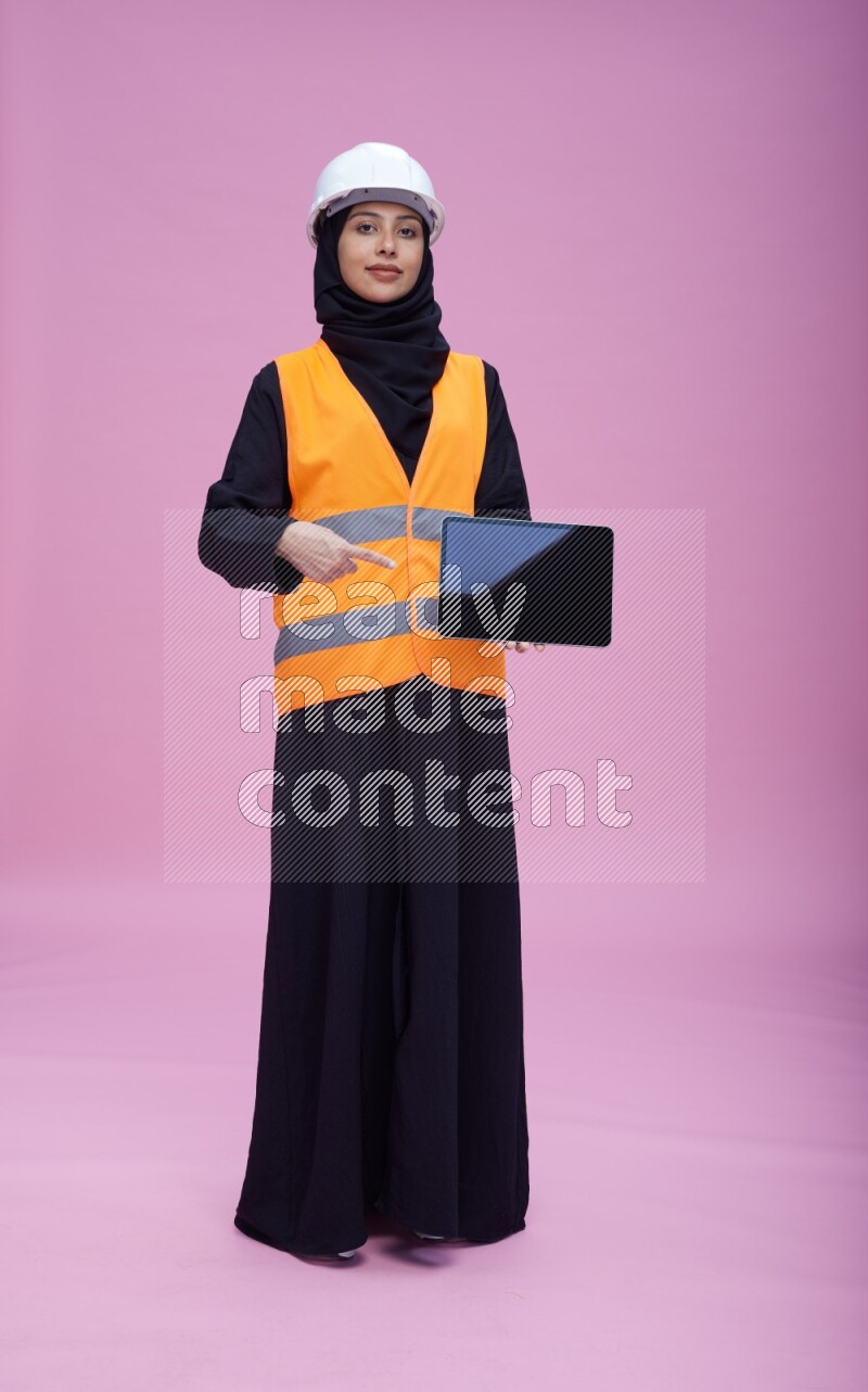 Saudi woman wearing Abaya with engineer vest and helmet standing showing tablet to camera on pink background