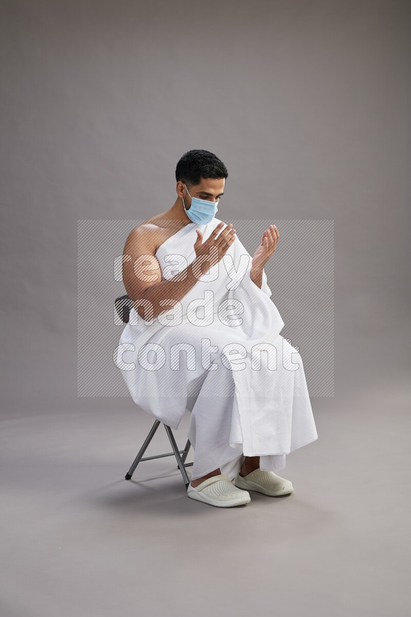 A man wearing Ehram with face mask sitting on chair dua'a on gray background