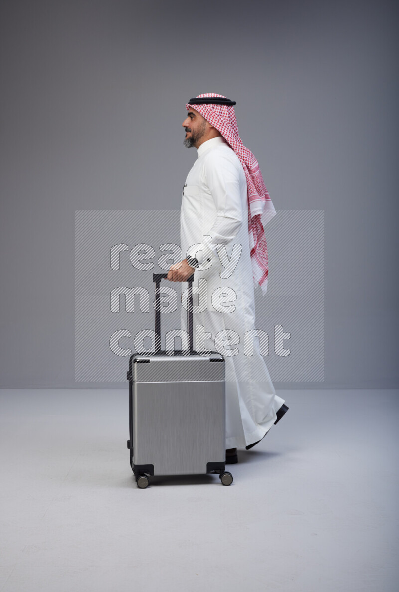 Saudi man wearing Thob and red Shomag standing holding Travel bag on Gray background