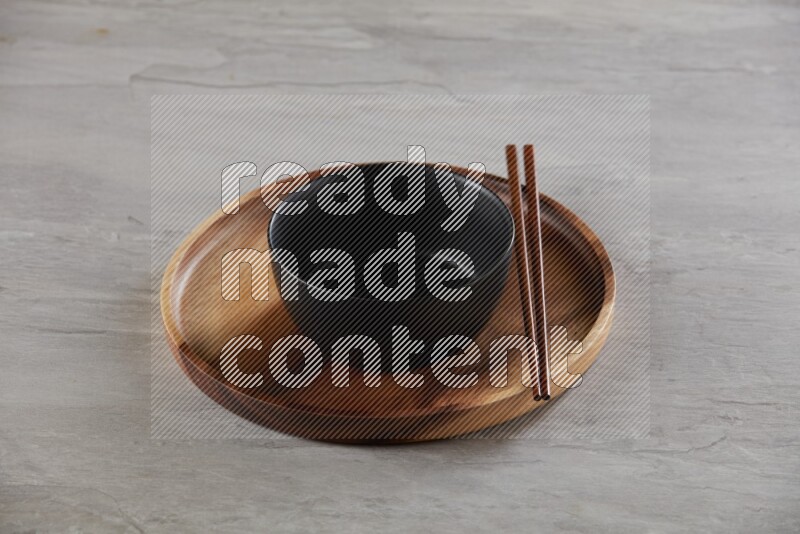 black ceramic round bowl on top of brown wood round plate and wood chopsticks, on grey textured countertop