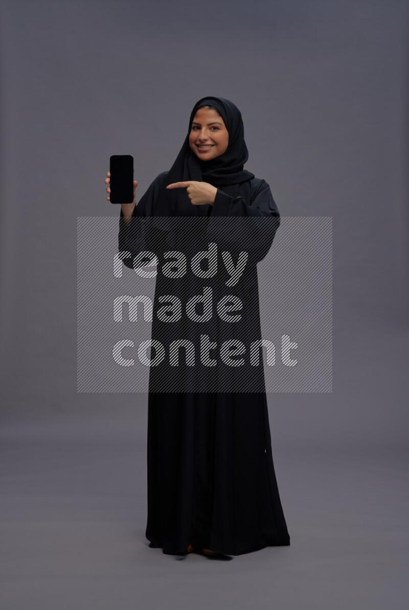 Saudi woman wearing Abaya standing showing phone to camera on gray background