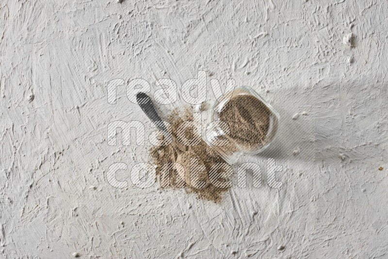 Flipped glass spice jar full of black pepper powder with a metal spoon full of it on textured white flooring