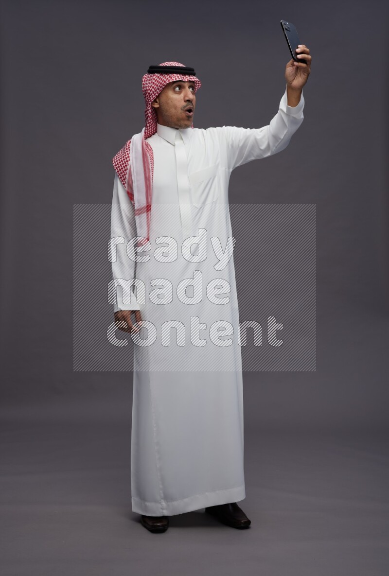 Saudi man wearing thob and shomag standing taking selfie on gray background
