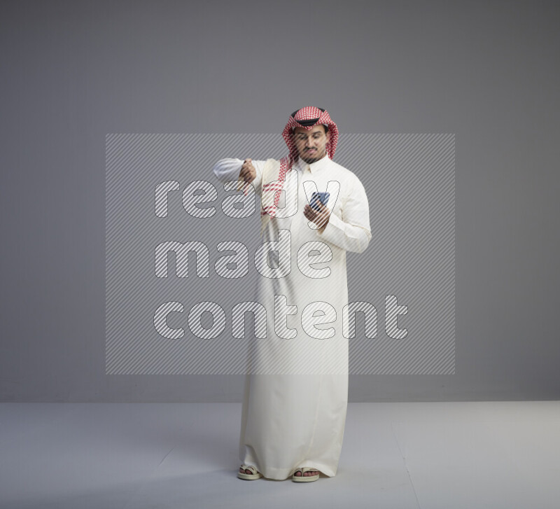 A  Saudi man standing wearing thob and red shomag texting on phone on gray background