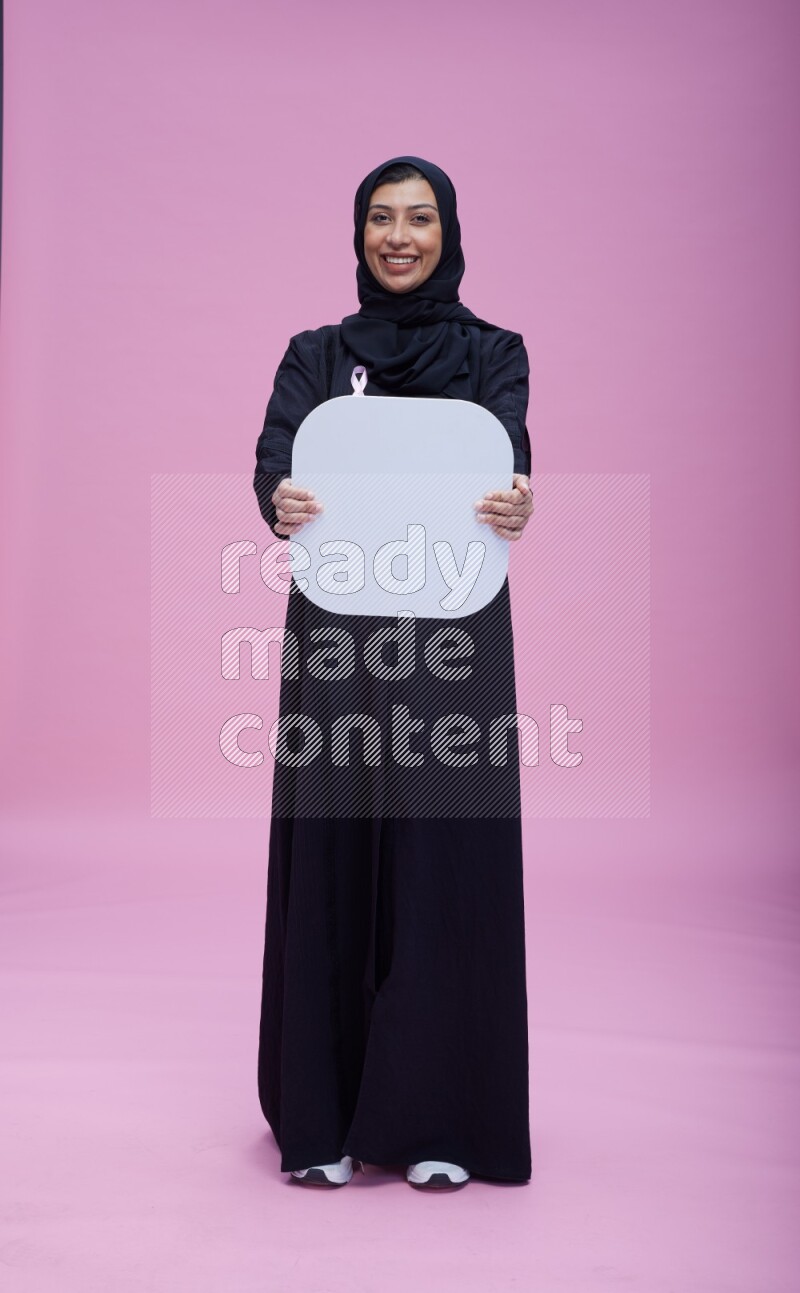 Saudi woman wearing pink ribbon on Abaya standing holding social media signs on pink background