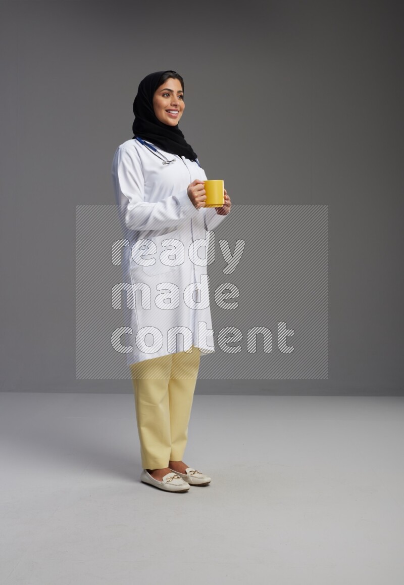 Saudi woman wearing lab coat with stethoscope standing holding mug on Gray background