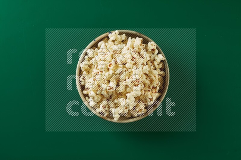 A beige ceramic bowl full of popcorn on a green background in a top view shot