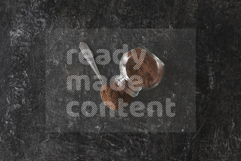 وعاء زجاجي للبهارات مقلوب وملعقة معدنية ممتلئان ببودرة القرنفل على أرضية سوداء