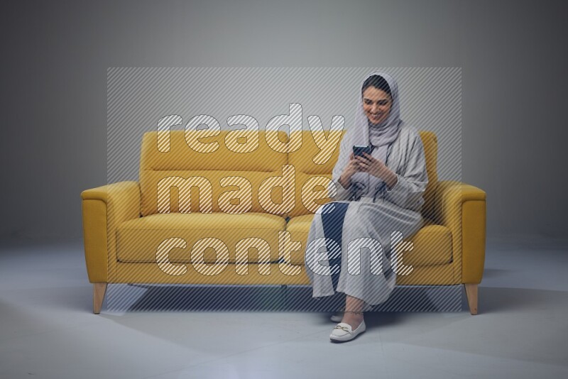 A Saudi woman wearing a light gray Abaya and white head scarf sitting on a yellow sofa and using her phone eye level on a grey background