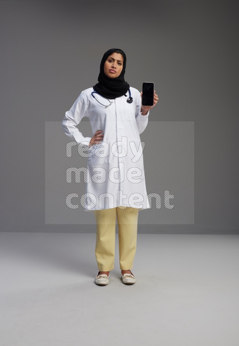 Saudi woman wearing lab coat with stethoscope standing showing phone to camera with sign in the back on Gray background