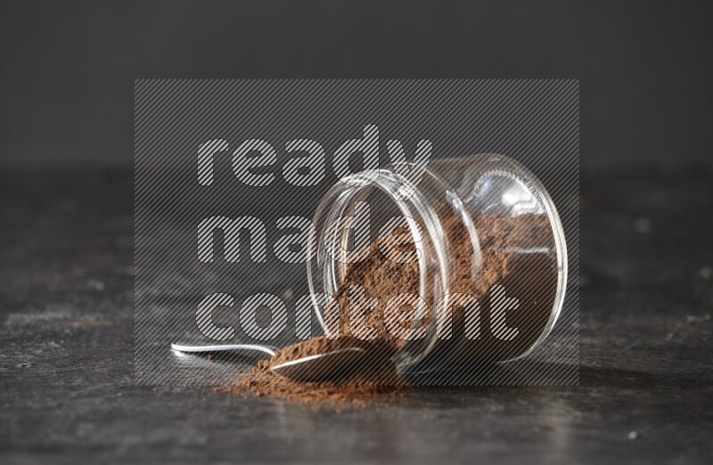 A flipped glass jar and metal spoon full of cloves powder with cloves spread on a textured black flooring