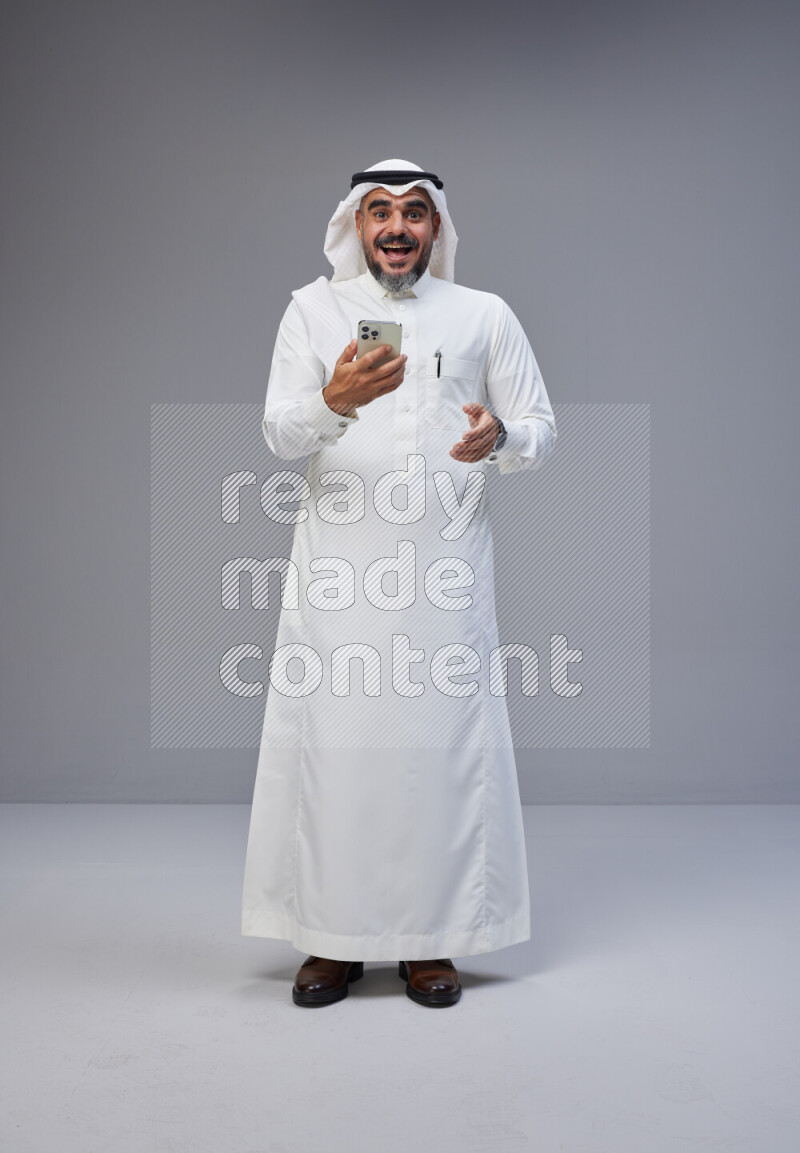 Saudi man Wearing Thob and white Shomag standing texting on phone on Gray background