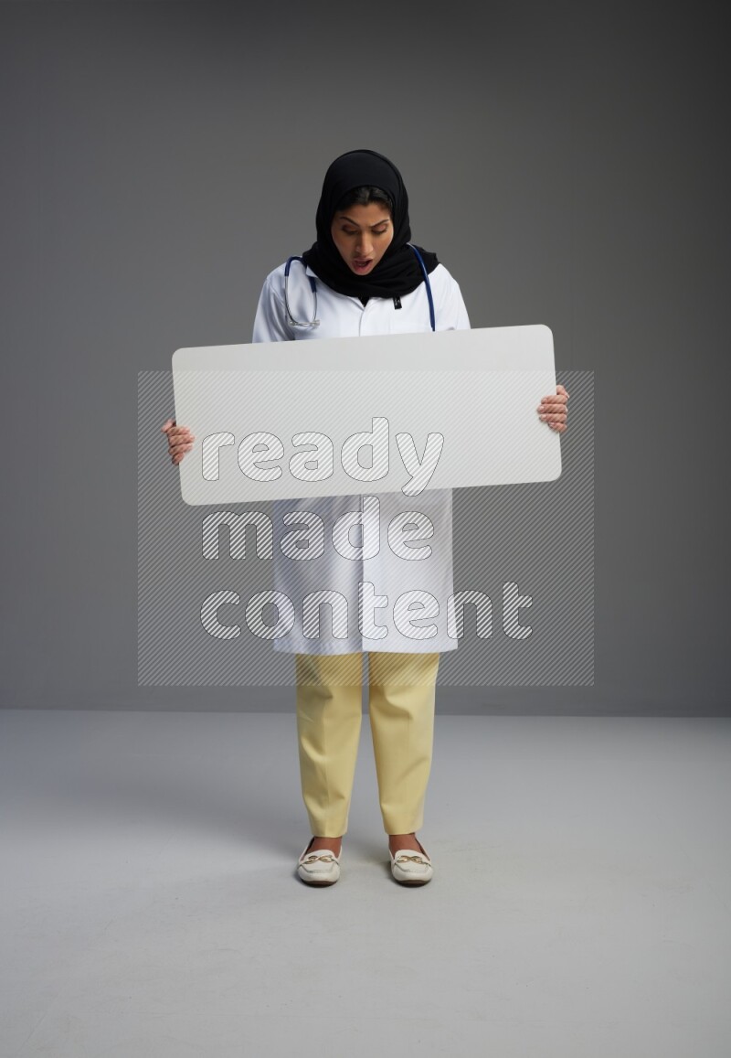 Saudi woman wearing lab coat with stethoscope standing holding board on Gray background