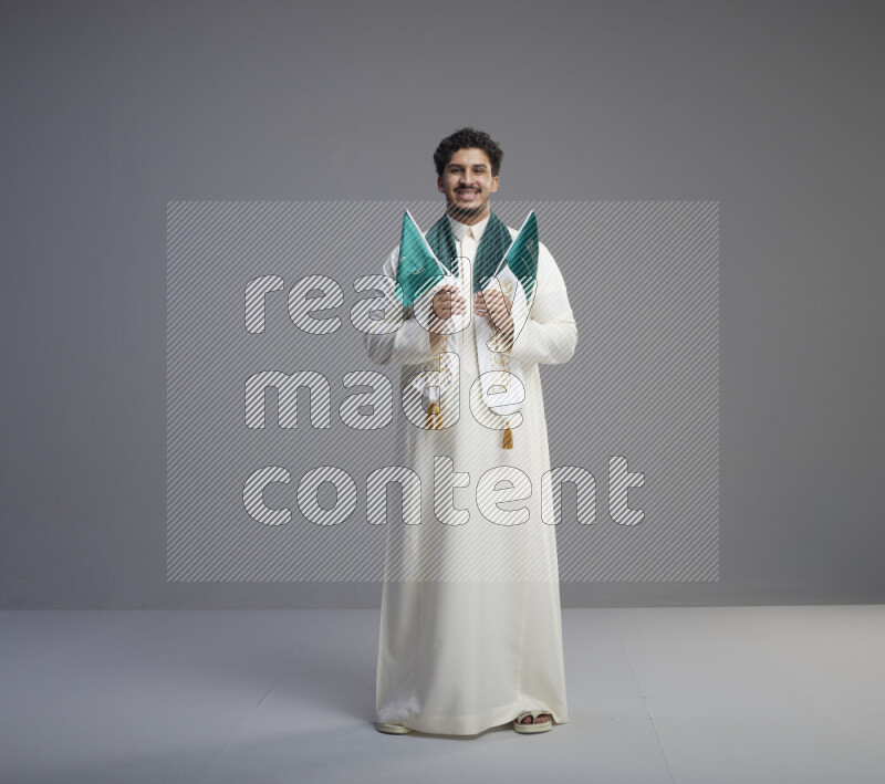 A Saudi man standing wearing thob and saudi flag scarf and holding small saudi flag on gray background