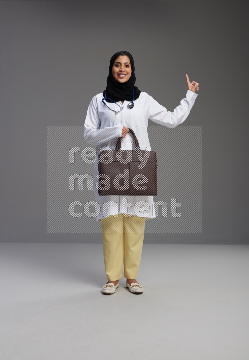 Saudi woman wearing lab coat with stethoscope standing holding bag on Gray background