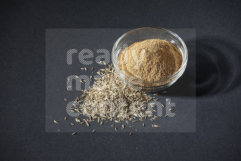 A glass bowl full of cumin powder with cumin seeds beside it on black flooring