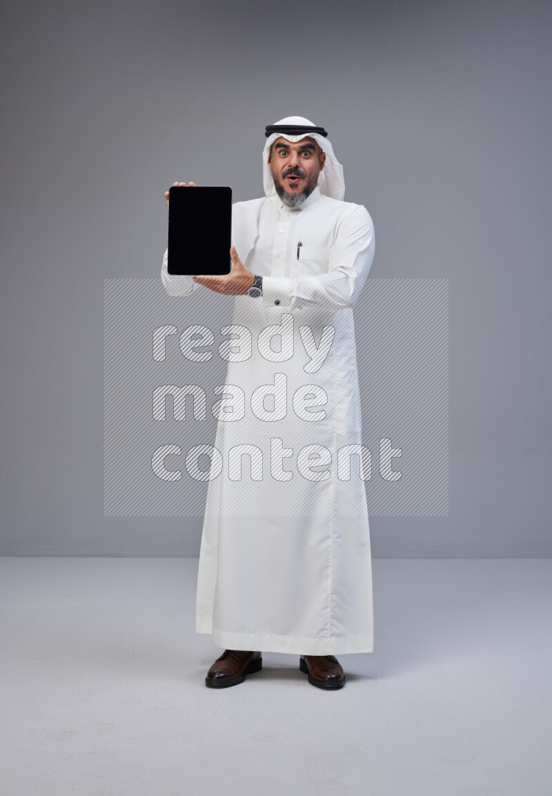 Saudi man Wearing Thob and white Shomag standing showing tablet to camera on Gray background