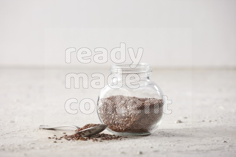 A glass spice jar full of flax seeds and a metal spoon full of the seeds on a textured white flooring