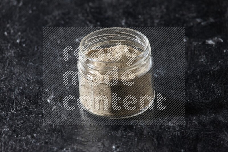A glass jar full of white pepper powder on textured black flooring