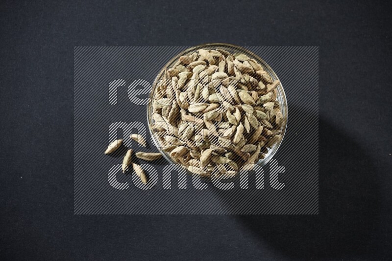 A glass bowl full of cardamom and more seeds spreaded beneath the bowl on black flooring