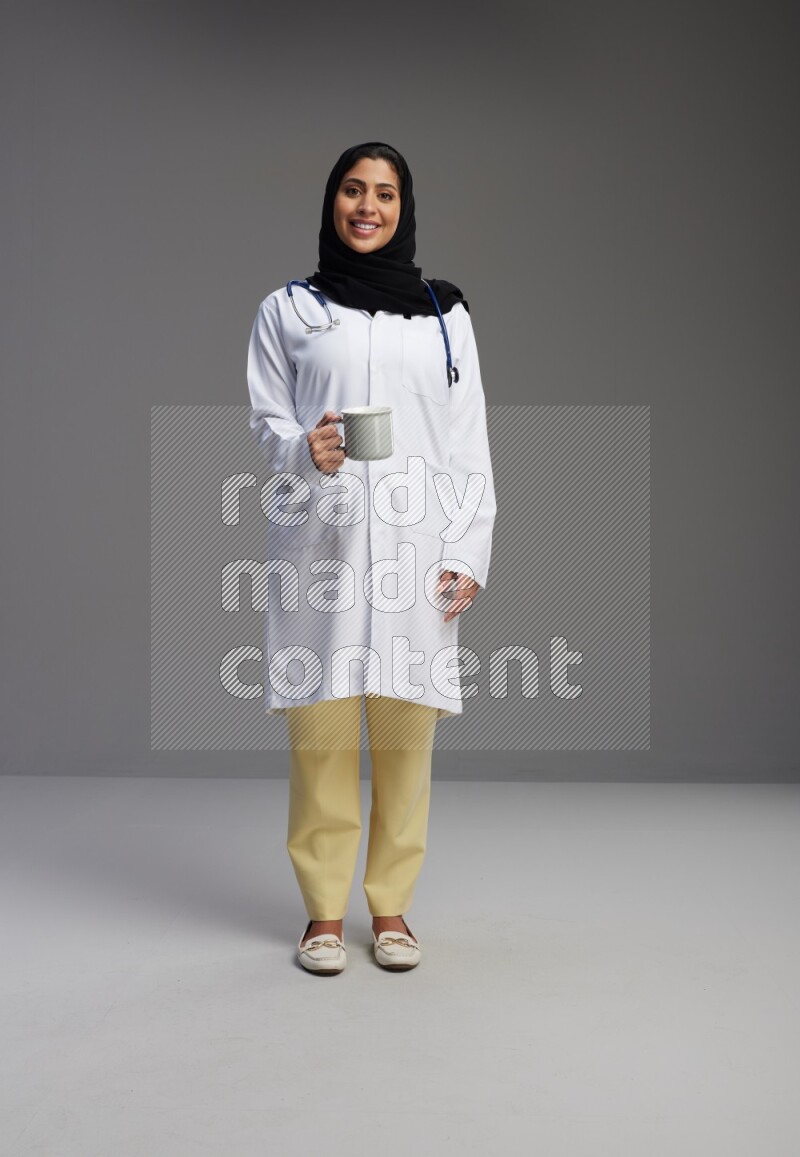 Saudi woman wearing lab coat with stethoscope standing holding mug on Gray background