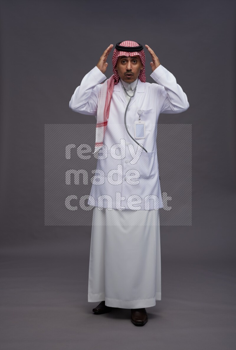 Saudi man wearing thob with lab coat and shomag with pocket employee badge with stethoscope standing interacting with the camera on gray background