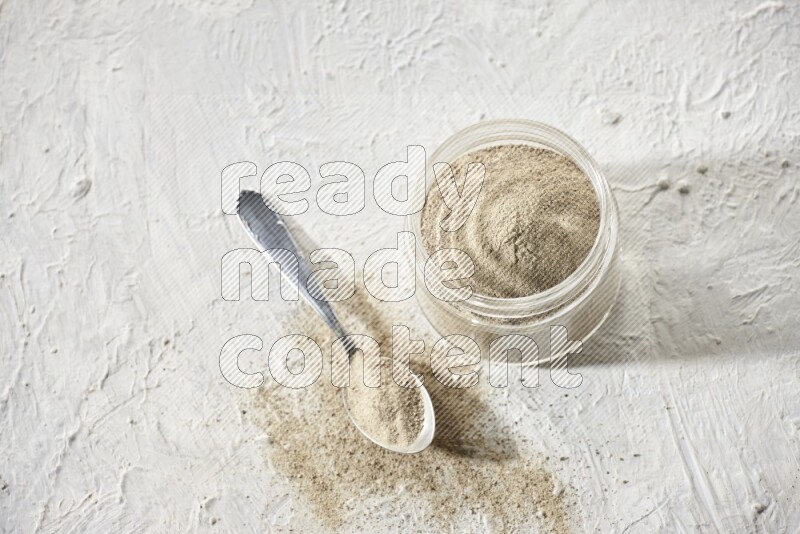 A glass jar and a metal spoon full of white pepper powder on textured white flooring