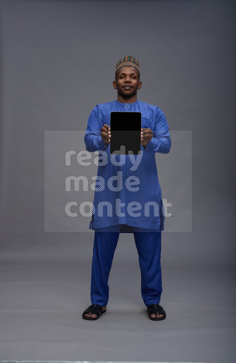 Man wearing Nigerian outfit standing showing tablet to camera on gray background