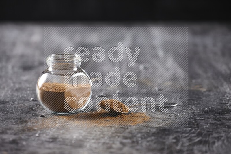 Herbal glass jar full of cinnamon powder and a metal spoon full of powder on textured black background