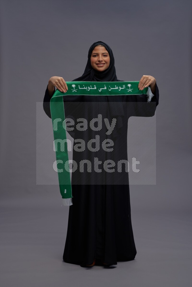 Saudi woman wearing Abaya standing holding Saudi flag on gray background