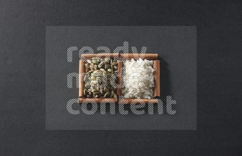 مربعان من أعواد القرفة ممتلئان بالملح الأبيض وحبوب الهيل علي خلفية سوداء