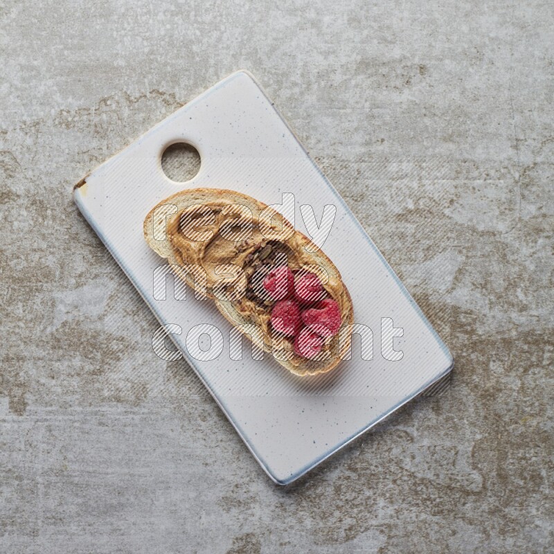 open faced peanut butter sandwich with raspberries and chocolate granula on a grey textured background