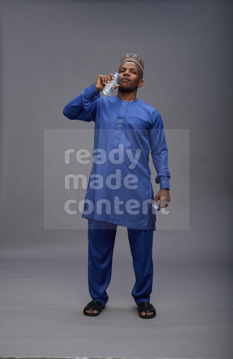 Man wearing Nigerian outfit standing drinking water on gray background