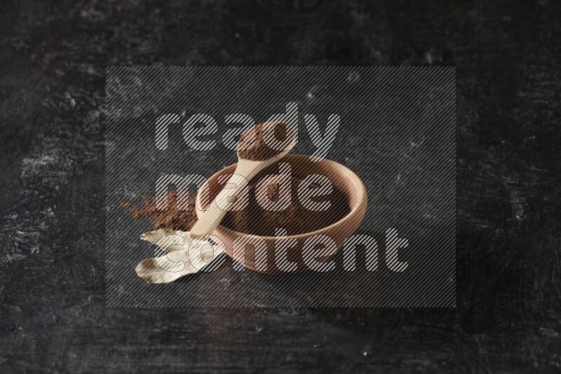 A wooden bowl and a wooden spoon full of cloves powder with laurel leaves on a textured black flooring