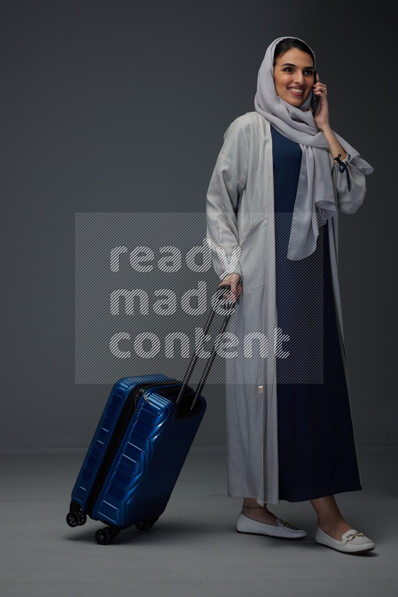 A Saudi woman wearing a light gray Abaya and head scarf standing and talking in the phone while holding shopping bags eye level on a grey background