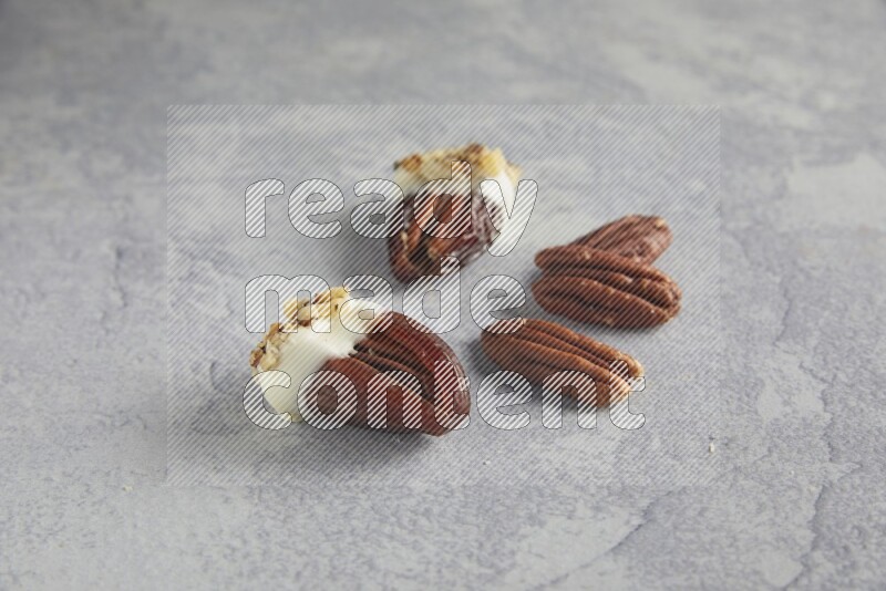 Two pecan stuffed dates covered with white chocolate and chopped walnuts with unroasted pecans on alight grey background