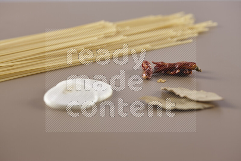 Raw pasta with different ingredients such as cherry tomatoes, garlic, onions, red chilis, black pepper, white pepper, bay laurel leaves, rosemary and cardamom on beige background