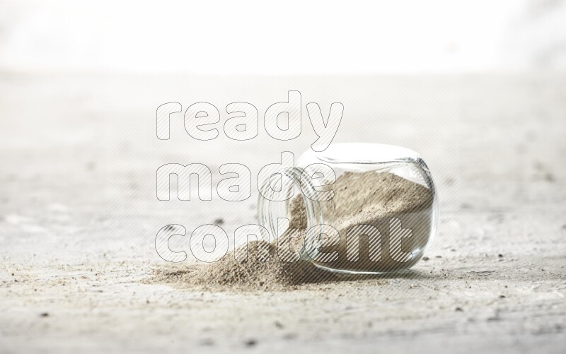 A flipped herbal glass jar full of white pepper powder with spilled powder on textured white flooring