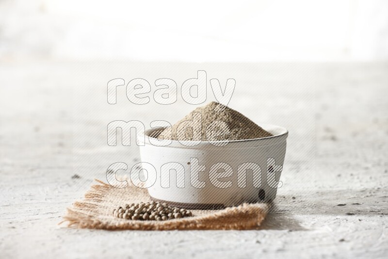 White pottery bowl full of white pepper powder set on a burlap piece of fabric with pepper beads and wooden pepper grinder on textured white flooring