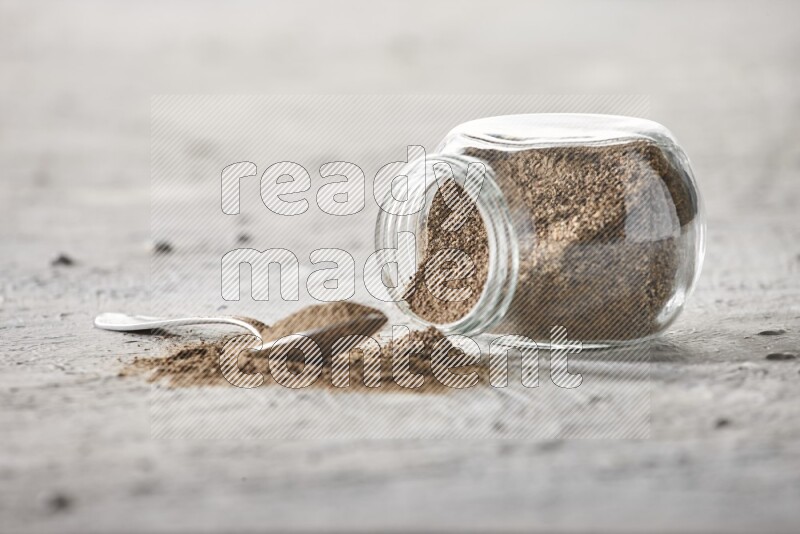 Flipped glass spice jar full of black pepper powder with a metal spoon full of it on textured white flooring