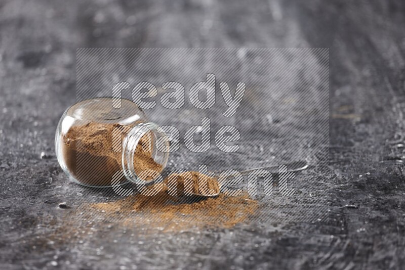 Herbal glass jar full of cinnamon powder flipped and a metal spoon on textured black background