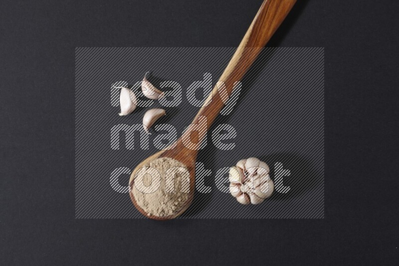 A wooden ladle full of garlic powder with a whole garlic bulb and some cloves beside it on a black flooring
